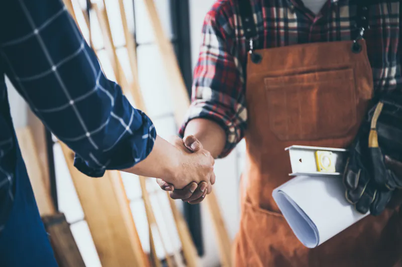 Handschlag zwischen einem Kunden und einem Tischler in Arbeitskleidung. Im Hintergrund ist Holz zu sehen. Symbolbild für eine erfolgreiche Partnerschaft bei Holz Disselnmeyer.
