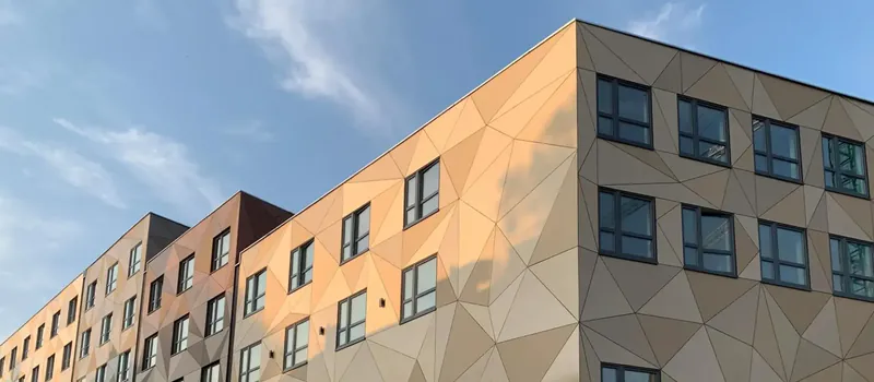 lumen-meteon-trespa-holz-disselnmeyer-f9b18bf5 Moderne Gebäudefassade mit geometrisch gemusterten Trespa Meteon Platten in Holzoptik bei einem Projekt von Disselnmeyer unter blauem Himmel.