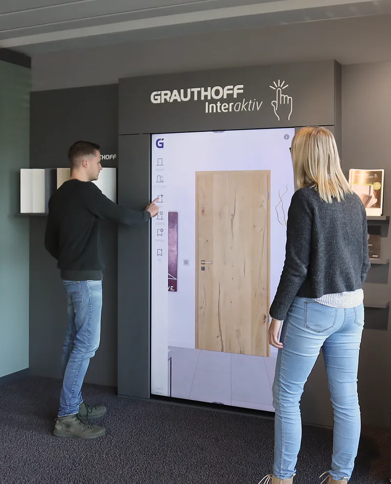 Zwei Personen konfigurieren interaktiv eine Holztür am digitalen Grauthoff-Terminal in einer Ausstellung.