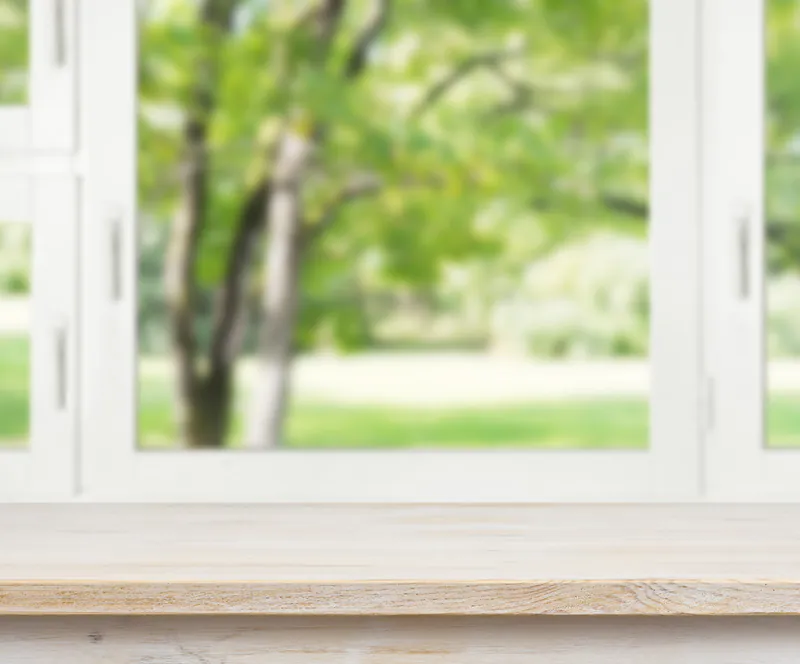 fensterbank-2204313a Helle Holzfensterbank vor einem Fenster mit Blick in den unscharfen, grünen Garten.