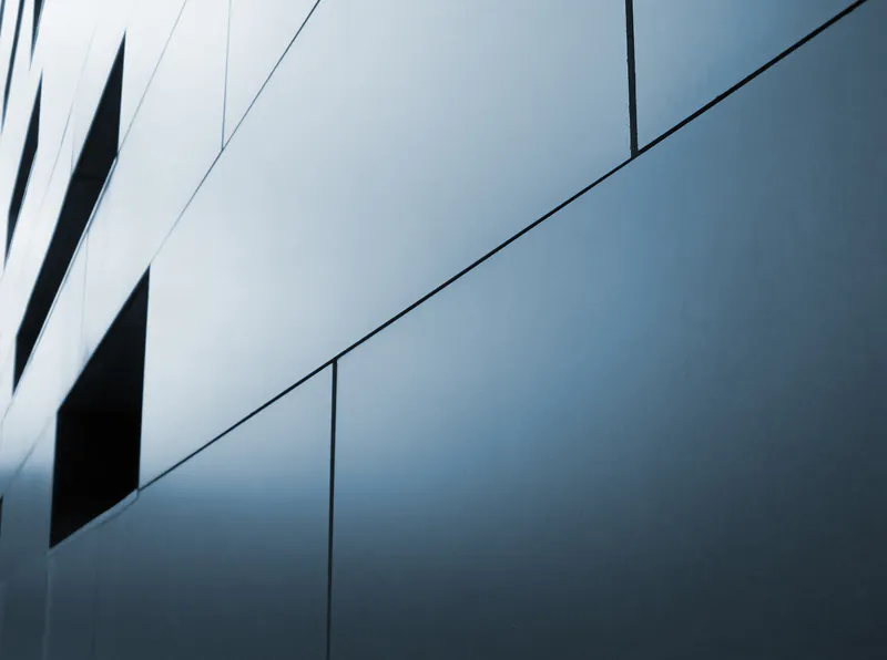 Moderne Fassade eines Gebäudes mit geometrischen Metallplatten und dunklen Fensteröffnungen in perspektivischer Ansicht.