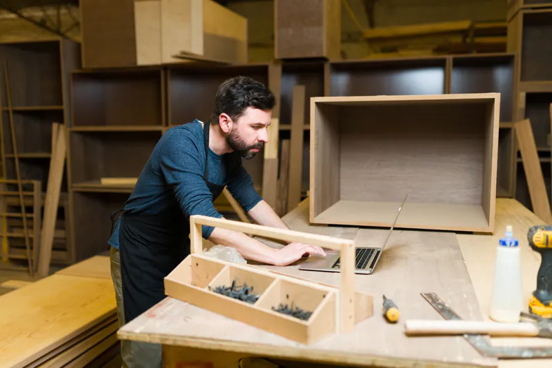 Ein Tischler bei der digitalen Planung von Möbeln am Laptop in seiner Holzwerkstatt. Eine gelungene Verbindung von traditionellem Handwerk und moderner Technik.